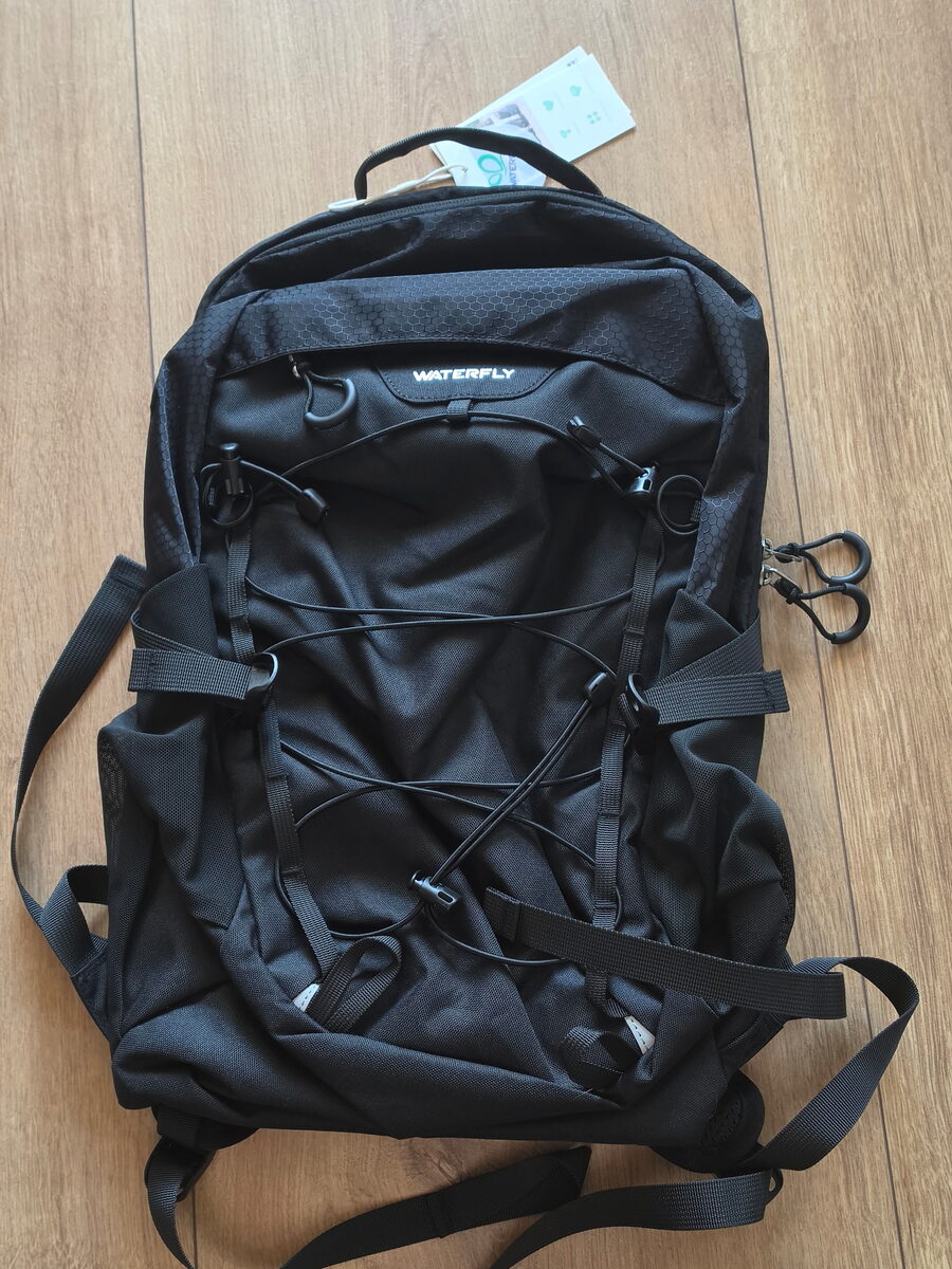 Front of the black WATERFLY 20L daypack laid flat, with its bungee cord and side straps